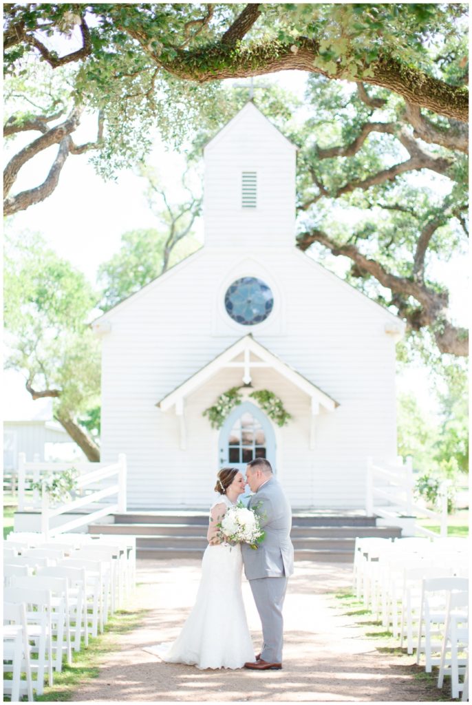 A bride and groom share an intimate moment in front of a white wedding chapel under Texas oak trees.