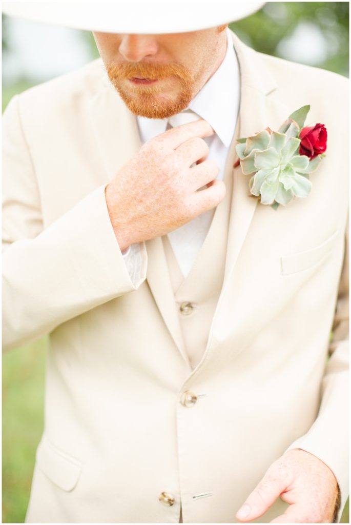 A groom adjusts his white tie while wearing a white cowboy hat and a succulent boutonniere. 