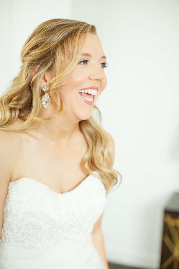 A bride wearing shiny dangling earrings laughs as she gets dressed for her wedding.