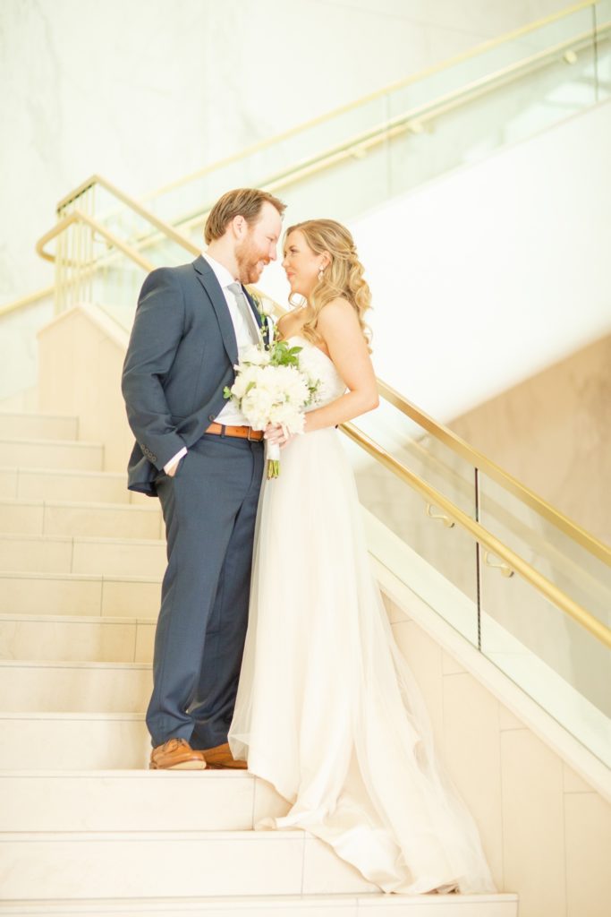 A bride and groom pose on a white marble stair case.