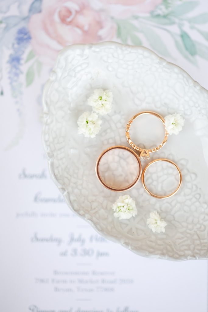 Three rose gold wedding rings are styled on a light blue dish over a blush and blue colored wedding invitation.