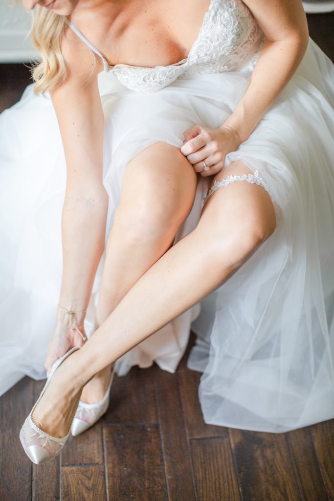 A bride puts on her shoes on her wedding day while wearing her wedding dress.