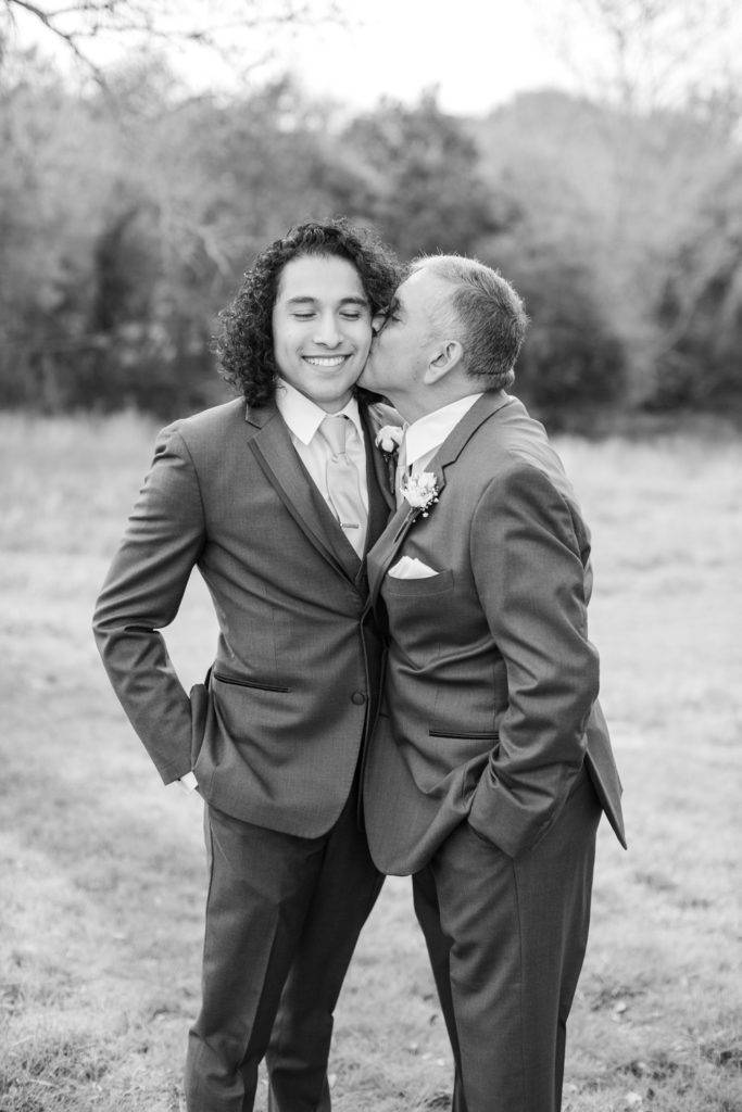 A groom's father gives the groom a kiss on the cheek on his wedding day.