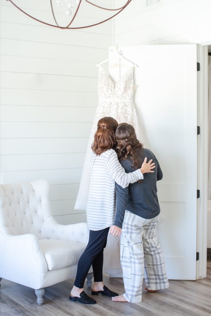 A mother and daughter admire the daughters wedding dress hung on a white door on the daughter's wedding day.