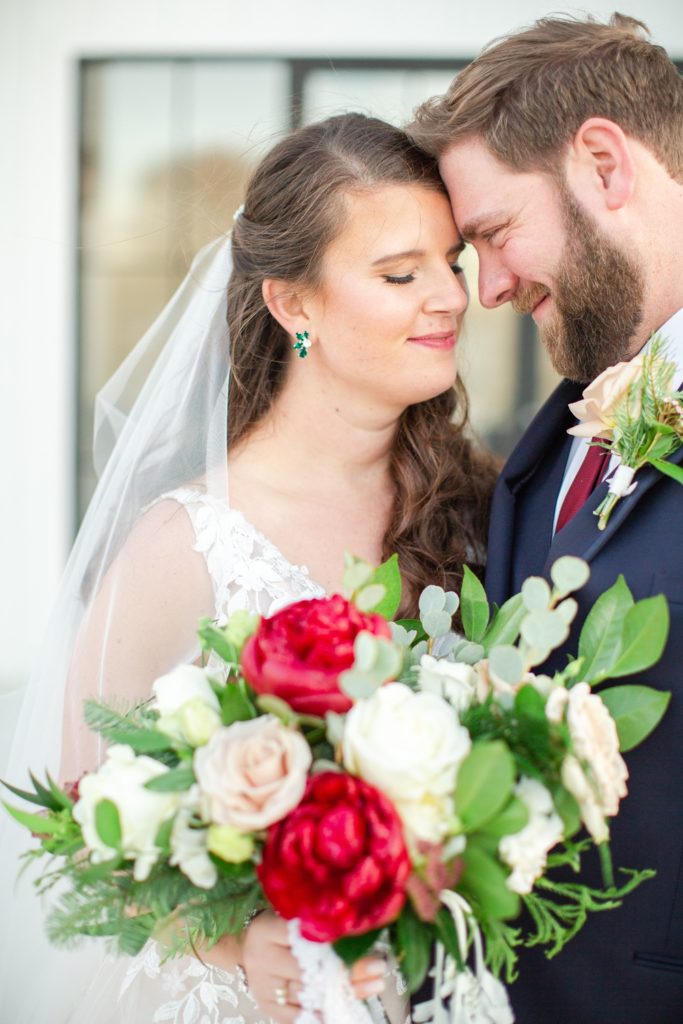 A bride and groom share a close moment huddled behind a winter red and green floral bouquet.
