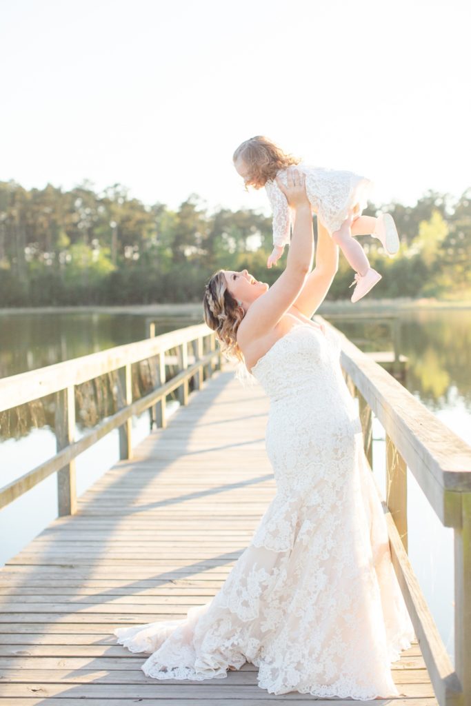 A bride, wearing her wedding dress, throws her flower girl into the air joyfully. The flower girl wears a white dress and pink keds.